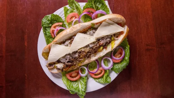 A cheesesteak on a long roll with melted provolone, plated with lettuce, tomato and onion.