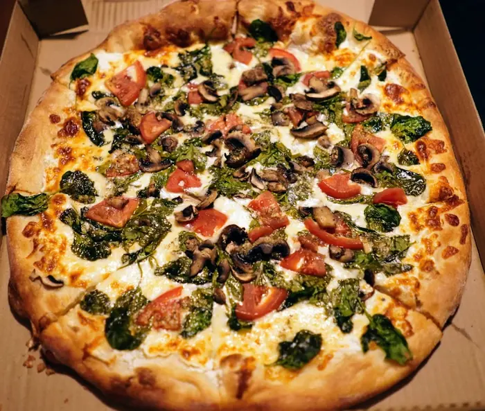 A white pizza with spinach, mushrooms, tomato and pesto, fresh from the oven in its box.